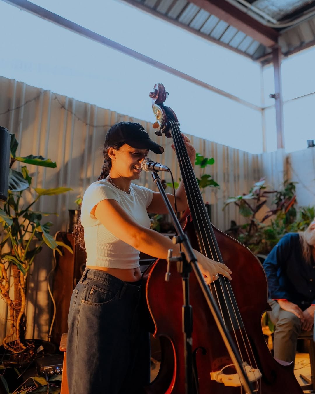 A young woman playing a double bass and singing into a microphone on a stage with plants in a warehouse space.