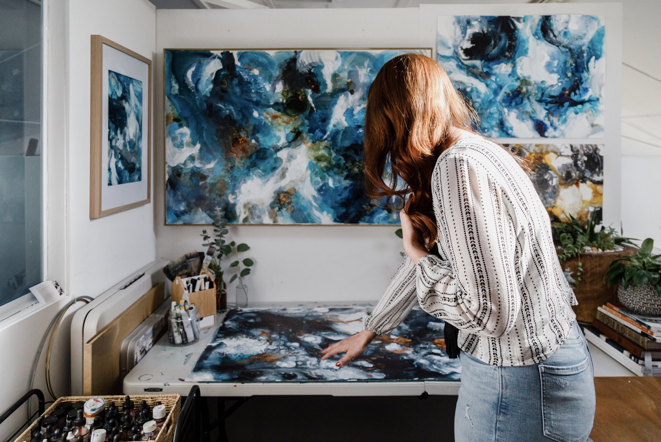 A woman with long red hair in a white patterned blouse and jeans is looking at and touching an abstract blue and white painting on a table inside an art studio.