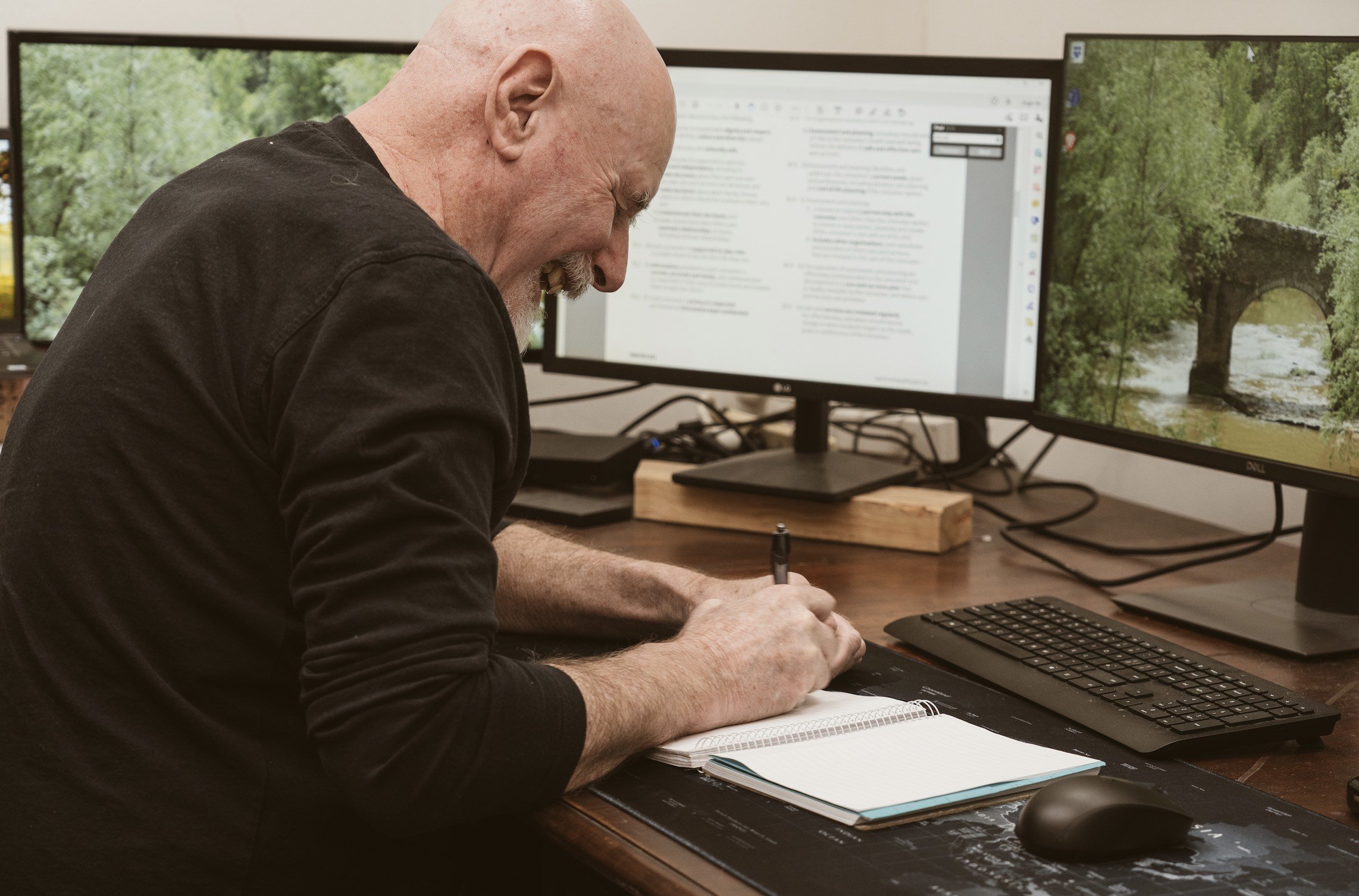 A man with a bald head and gray beard writing in a notebook at a desk with dual monitors, one displaying a document and the other a nature scene.