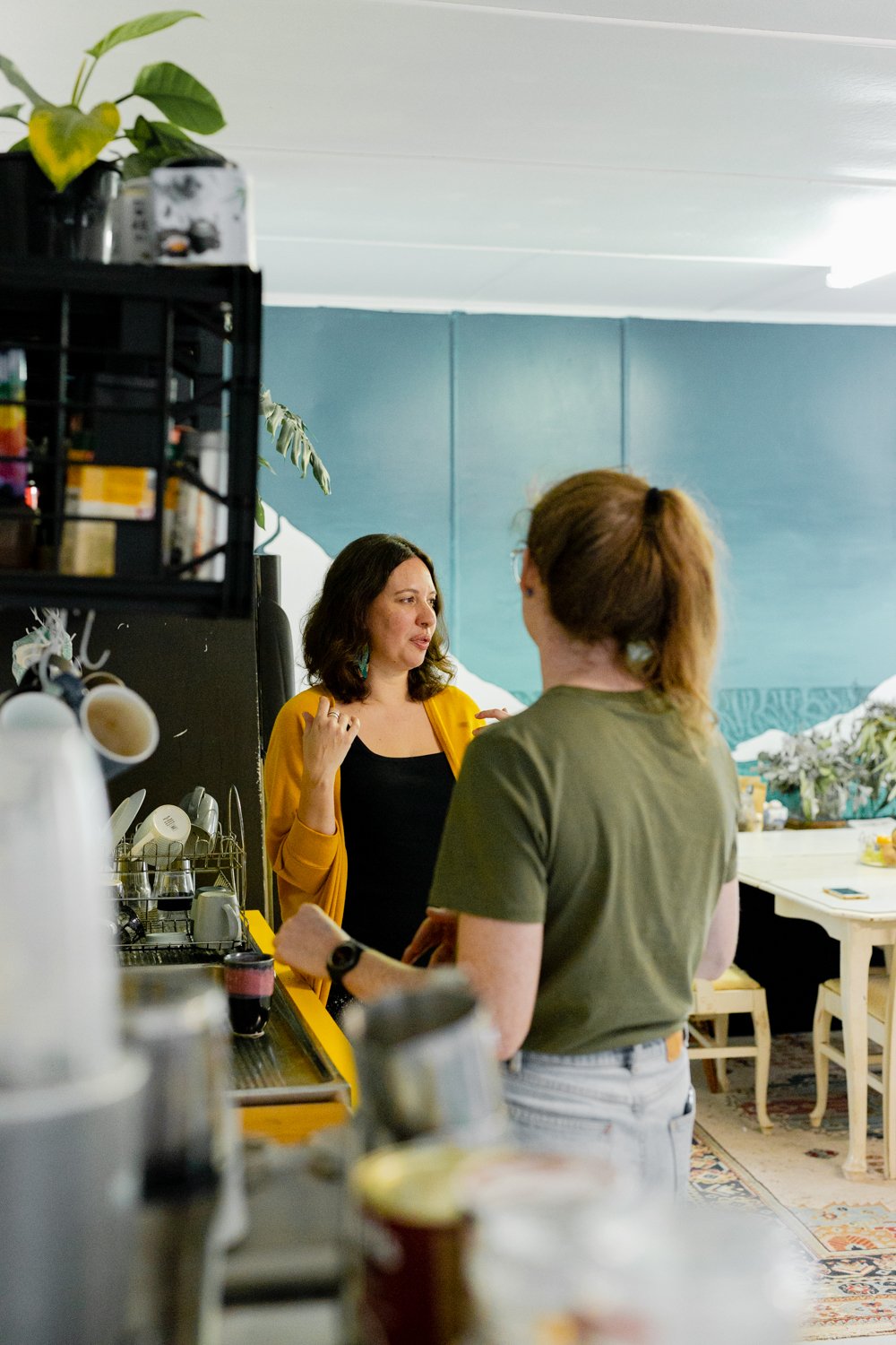 Two women having a conversation in a coffee shop, with kitchen equipment and a blue wall in the background.