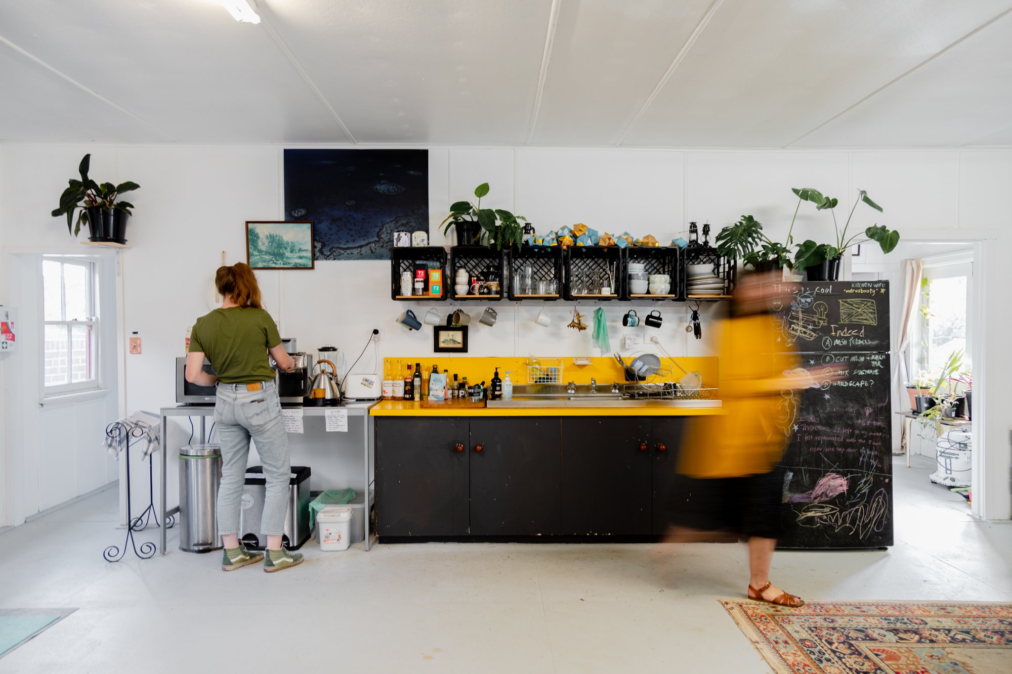 Two women in a modern, minimalist kitchen with yellow and black cabinets. One woman in a green top and grey pants stands at the counter, preparing food, while the other woman in a yellow shirt and black skirt walks past, blurred due to movement.