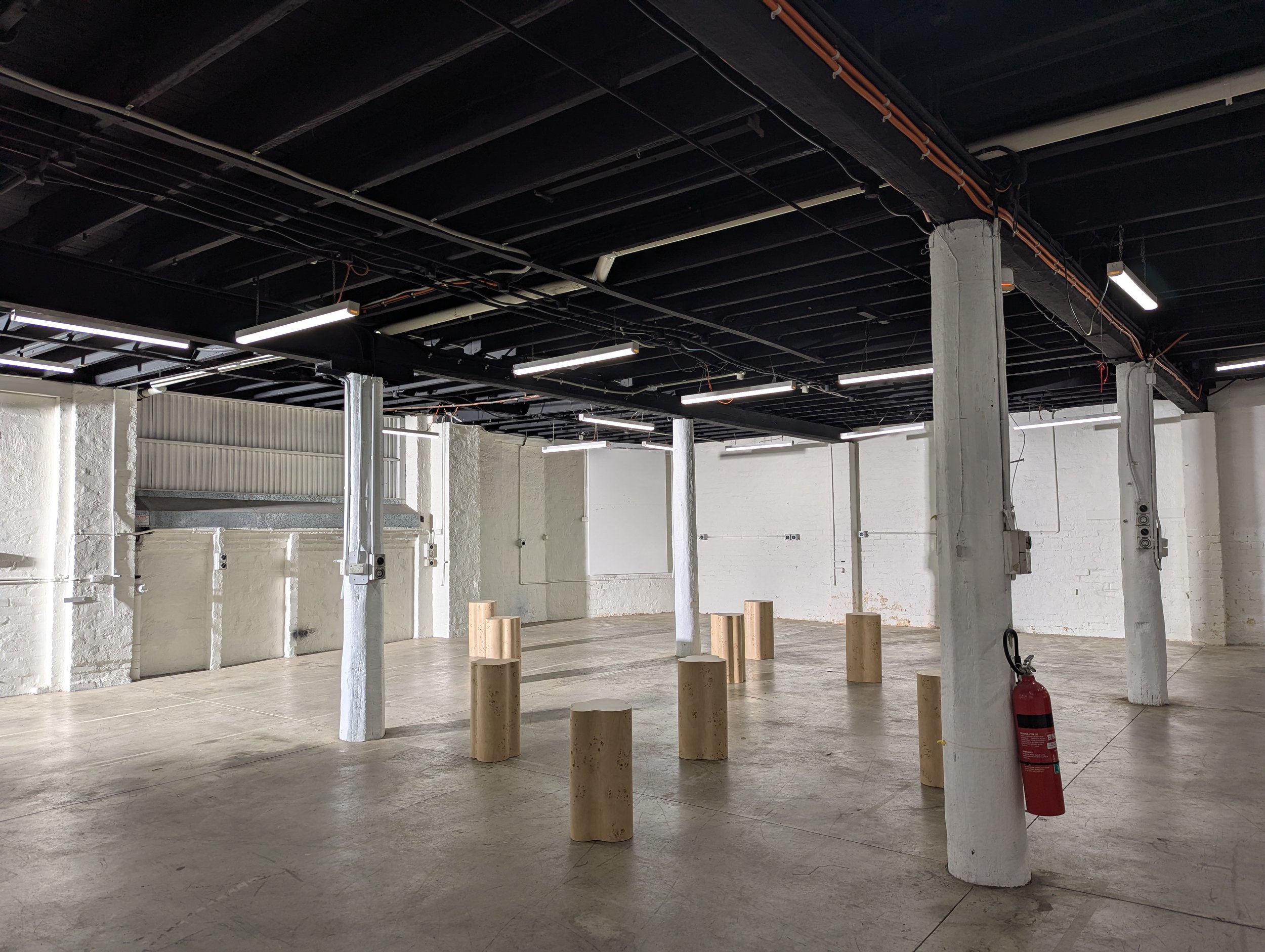 Empty industrial space with white painted brick walls, concrete floor, exposed black ceiling, white concrete columns, and wooden cylinders arranged on the floor for a photoshoot and film shoot.
